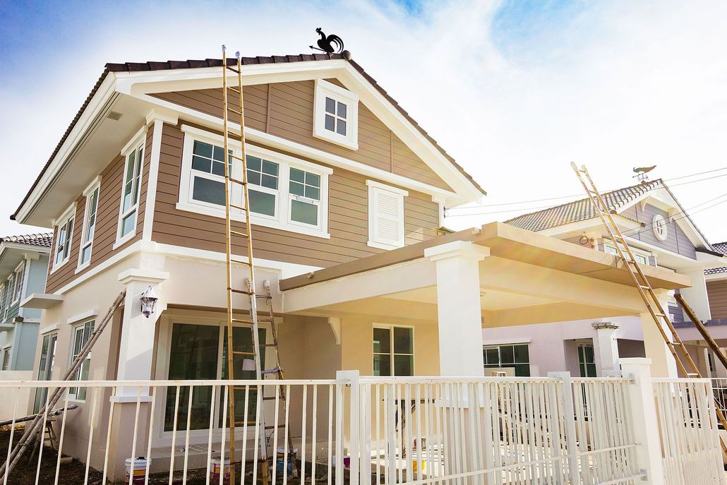 Two-story house with tan siding, white trim, and ladders on the exterior, behind a white fence.