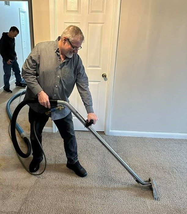 Man vacuuming a carpet, holding a wand. Another man in background. Neutral-colored room.