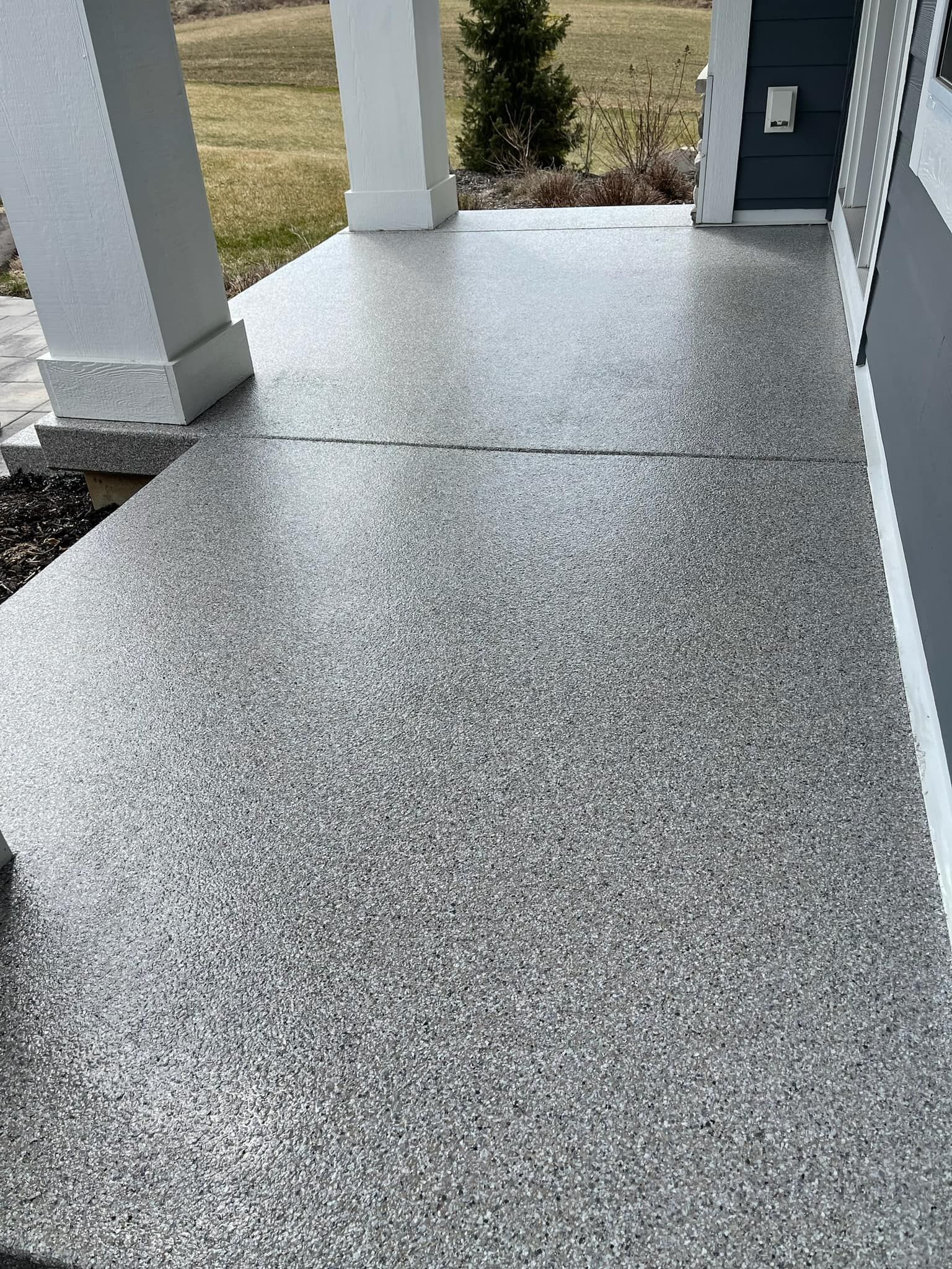 Concrete porch walkway with speckled gray flooring and white columns leading to an exterior door