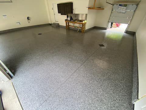 Empty garage with gray speckled floor, cabinets, workbench, and an open door letting in daylight