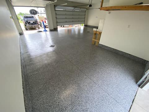 Empty garage with gray speckled floor, open door, and a white wall with a small stool.