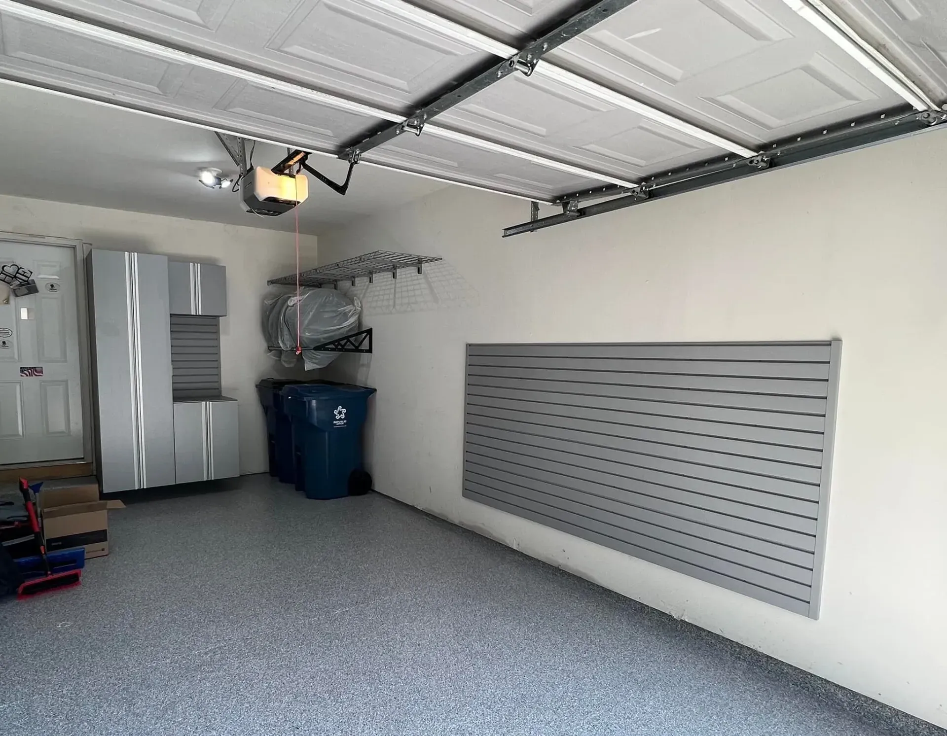 Empty garage with speckled floor, closed door, blue recycling bin, and wall-mounted storage rack