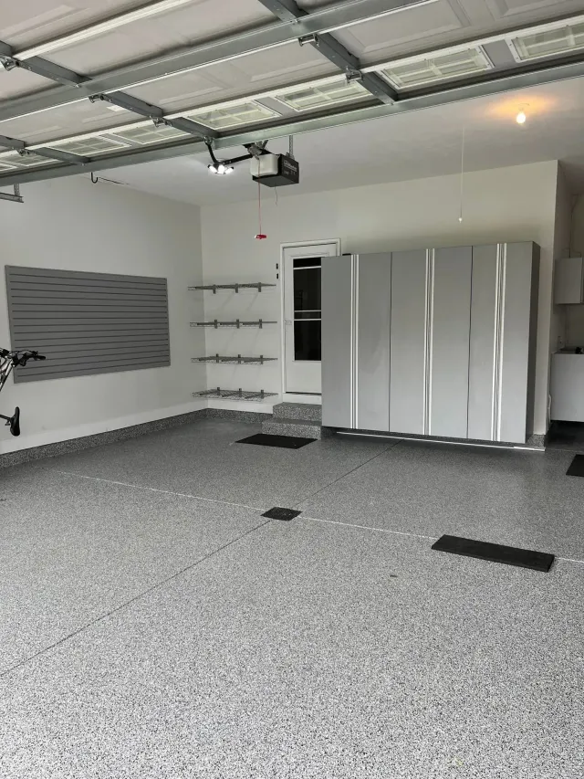 Empty garage with gray speckled floor, metal cabinets, wall shelves, and an overhead garage door opener.