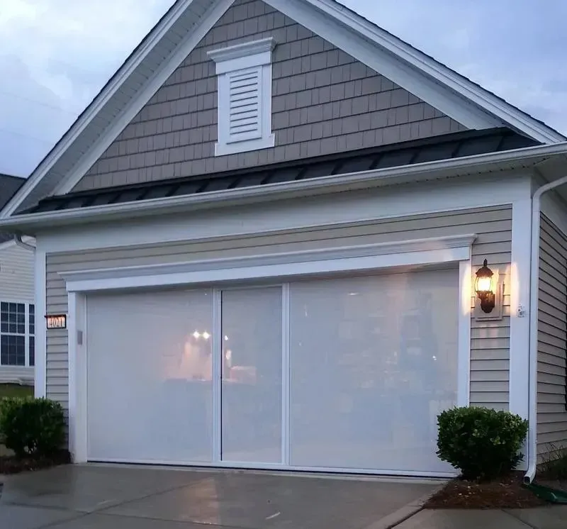 Gray suburban house with white garage doors, shingle siding, and a lit wall lantern at dusk