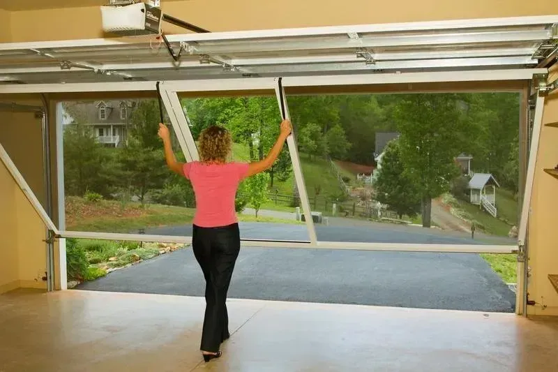 Person in a pink shirt opening a garage door, revealing a driveway and green neighborhood outside