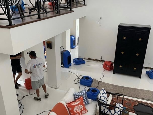 Two people in a bright living room near a staircase, surrounded by scattered cleaning supplies and furniture.