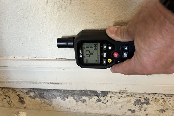 Handheld digital meter measuring a wall surface near a baseboard and floor.