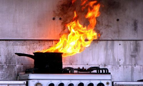 Kitchen stove with a pan on fire, bright flames rising against a gray wall
