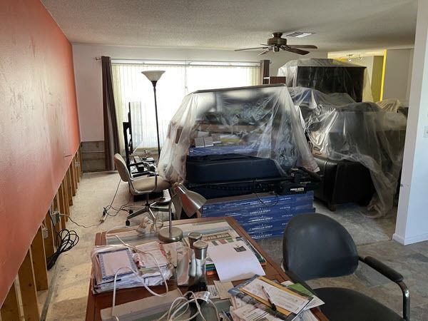 Cluttered living room with furniture wrapped in plastic during renovation, boxes, cords, and a floor lamp.