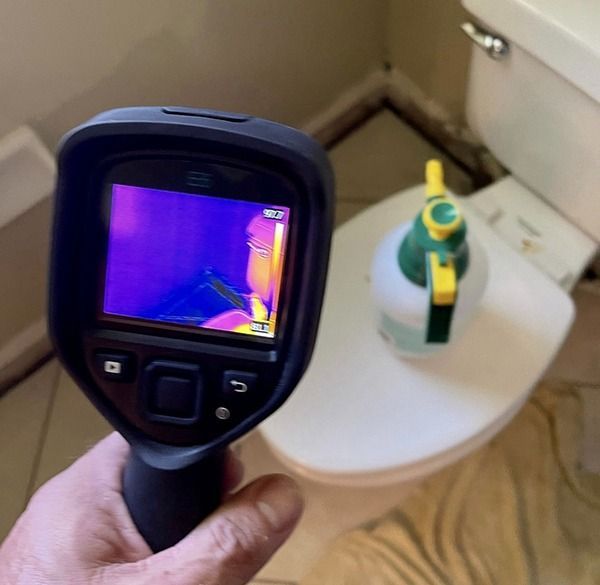 Handheld thermal camera aimed at a toilet with a green bottle on the seat in a bathroom.