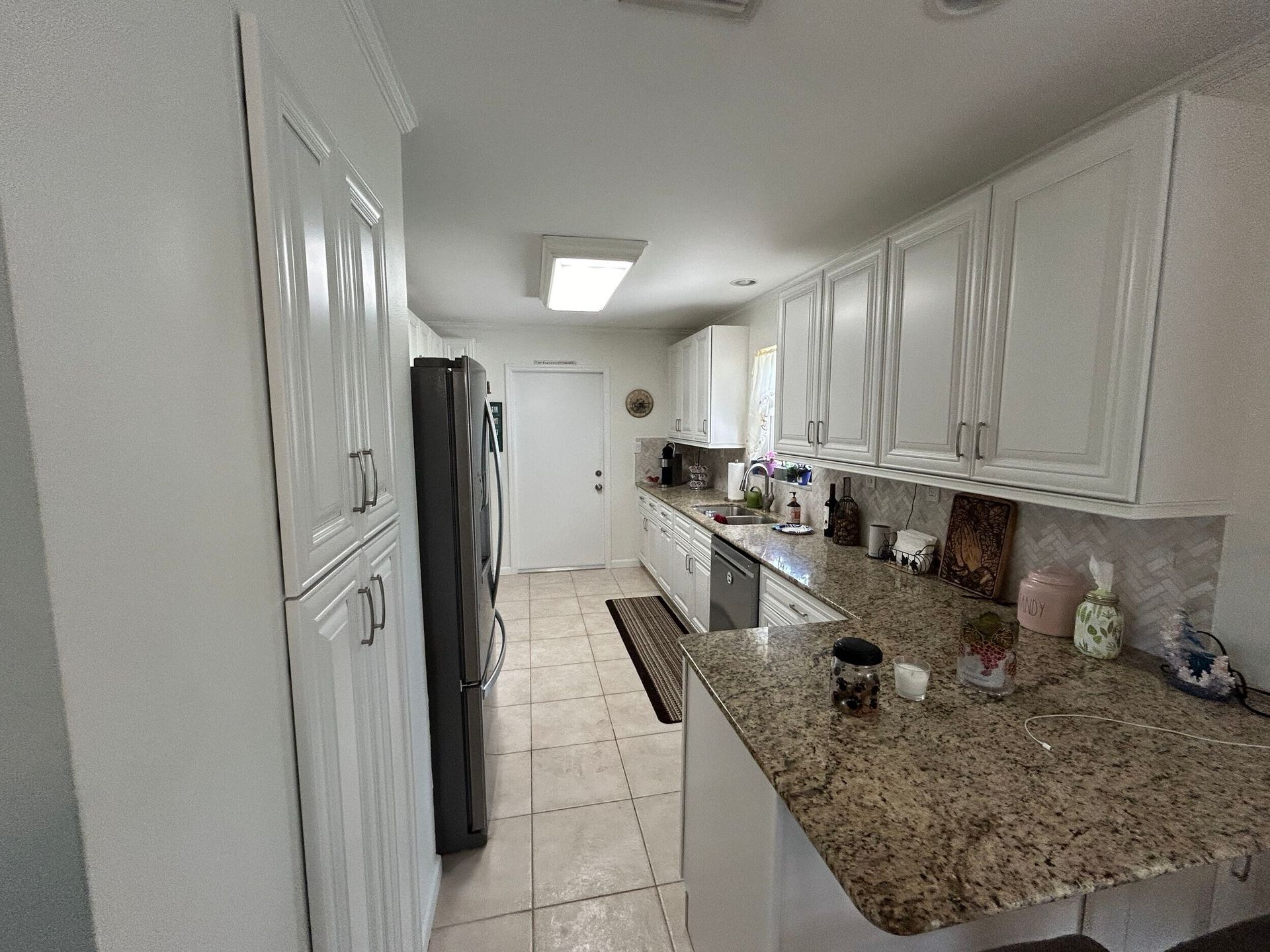Bright narrow kitchen with white cabinets, granite countertops, stainless fridge, and tiled floor.
