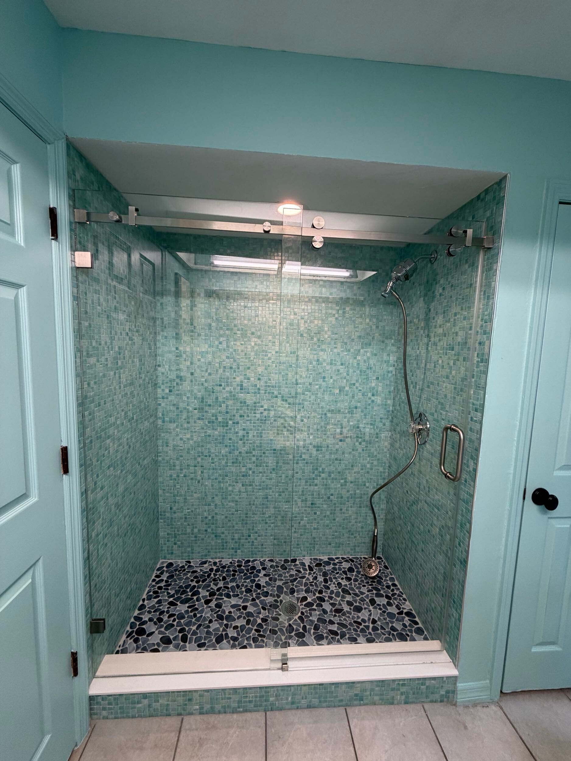 Empty teal-tiled shower stall with overhead light and black-and-white pebble floor.