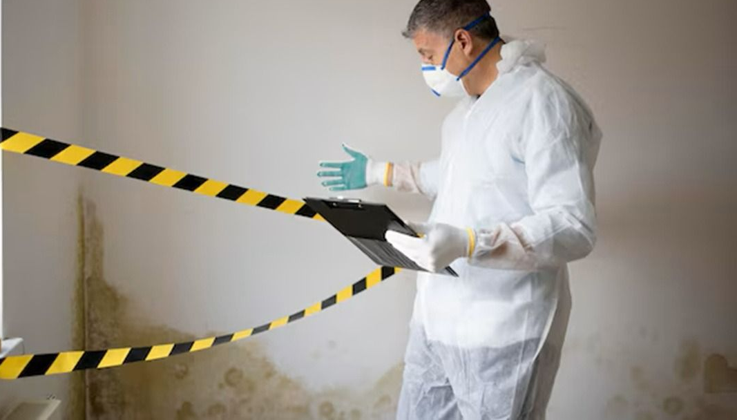 Person in protective coveralls inspecting a wall with hazard tape and a clipboard in a damaged room