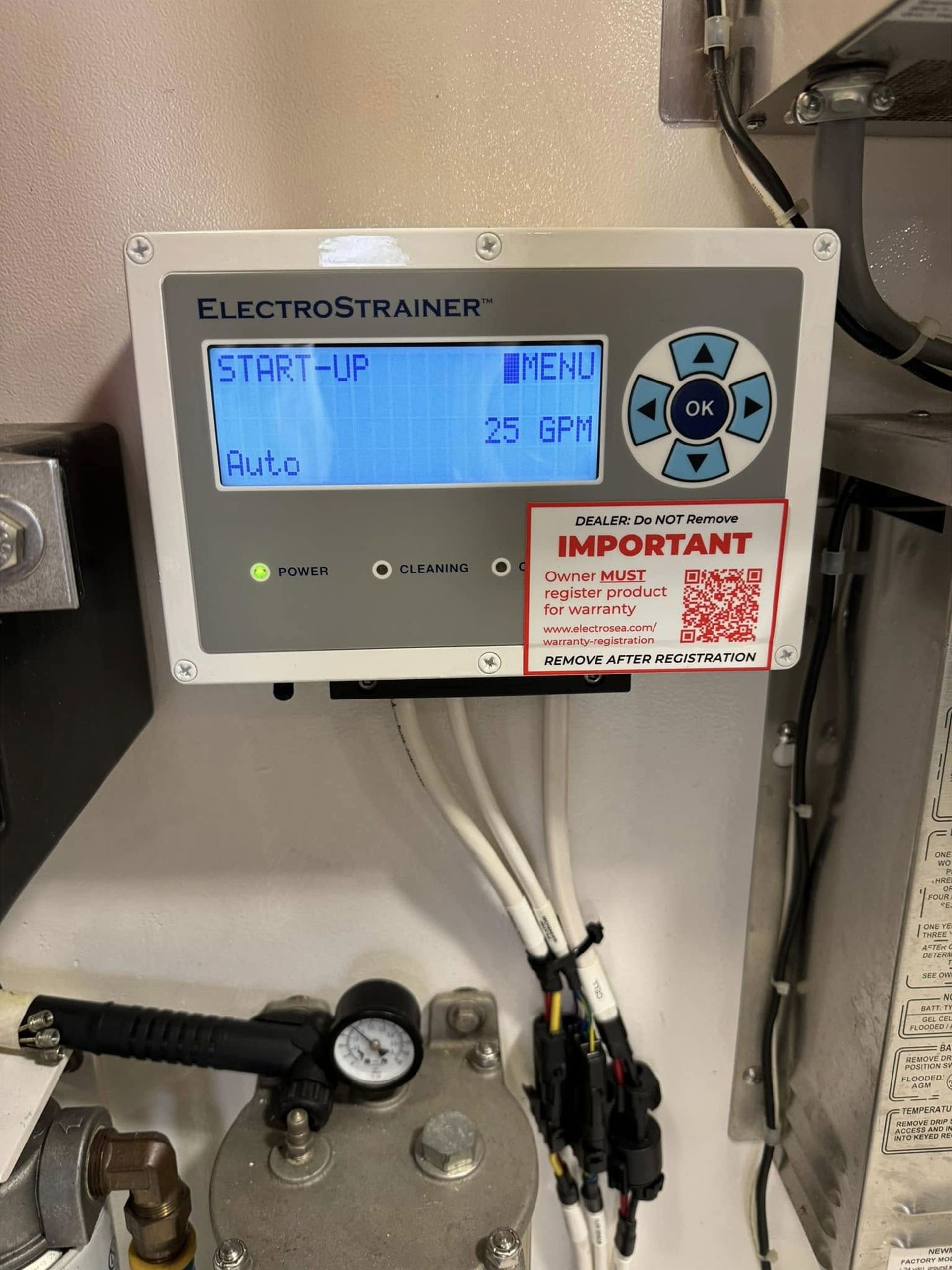 Wall-mounted boiler controller with blue display and warning label, above pipes and a pressure gauge