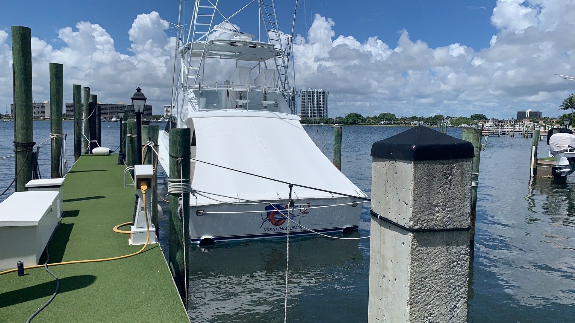 White boat docked at a marina beside green turf and pilings under a blue, partly cloudy sky