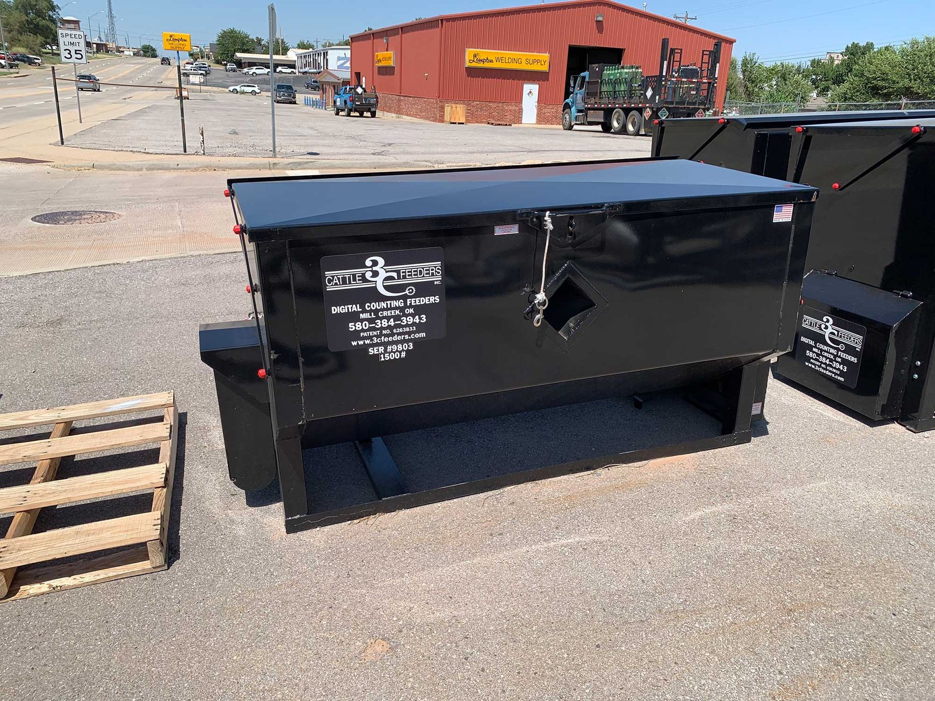 A large black dumpster is sitting in a parking lot next to a wooden pallet.
