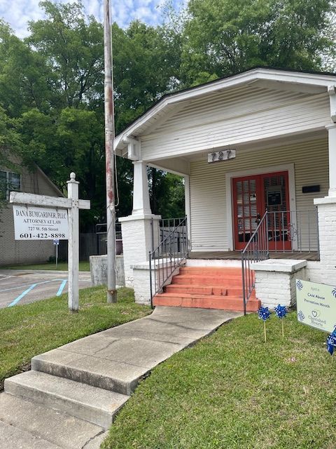 A white, one-story building with a red-framed entrance, a porch, and a sign on a pole, situated next to a sidewalk.