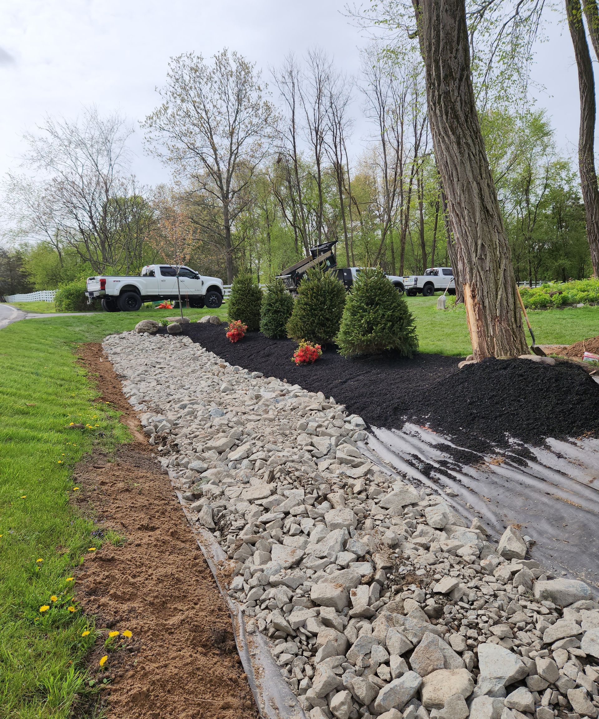 A garden bed with rocks, mulch, shrubs, and red/orange flowers. Trucks are parked in the background.