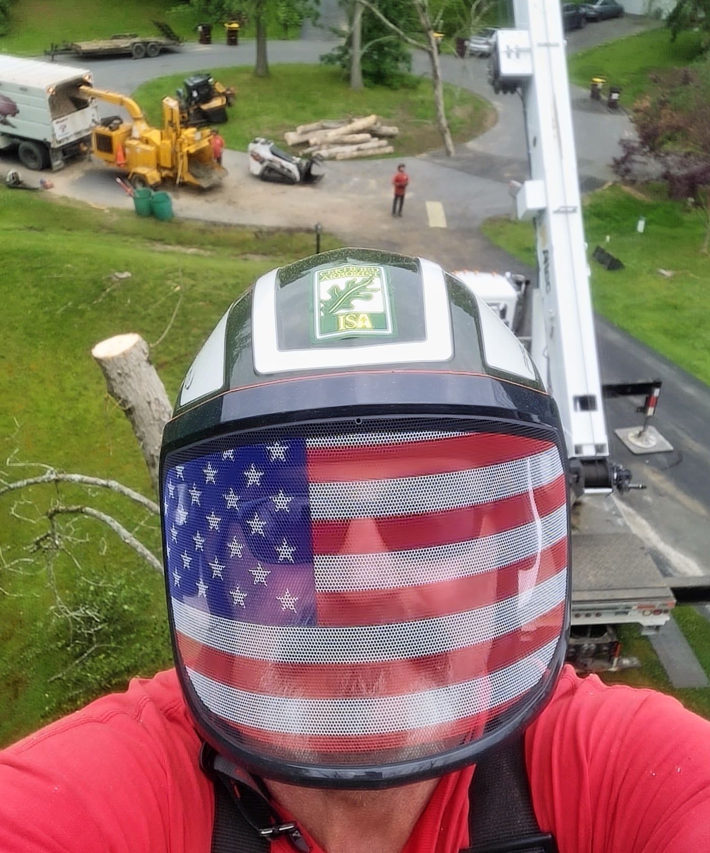 Person wearing a helmet with an American flag visor, tree trimming. Other workers and equipment are in the background.