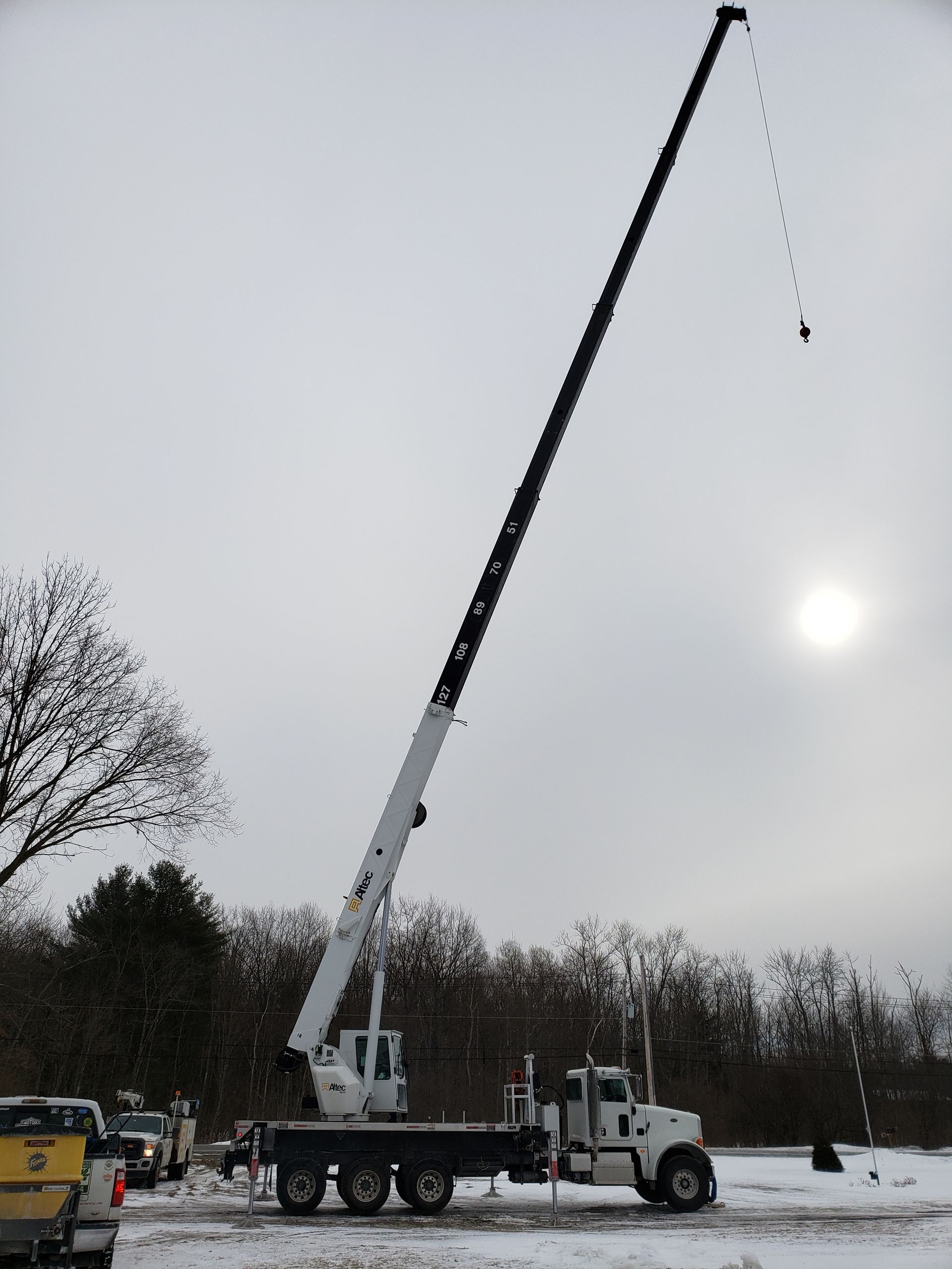 Crane truck with extended boom lifting something, in a snowy field with overcast sky.