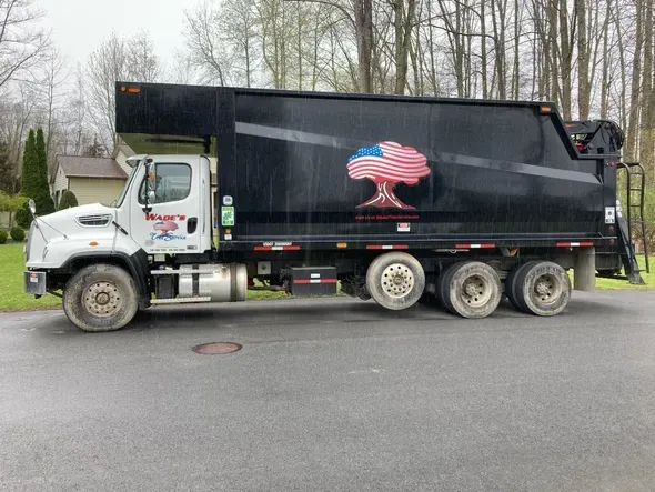Black garbage truck with an American flag tree logo on a residential street.