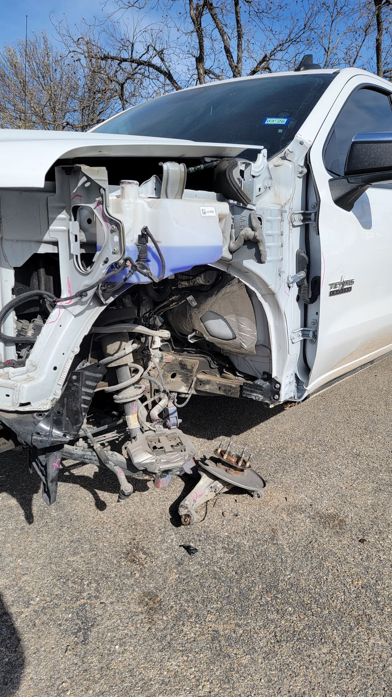 Severely damaged white pickup truck with missing front end, parked outdoors on gravel.