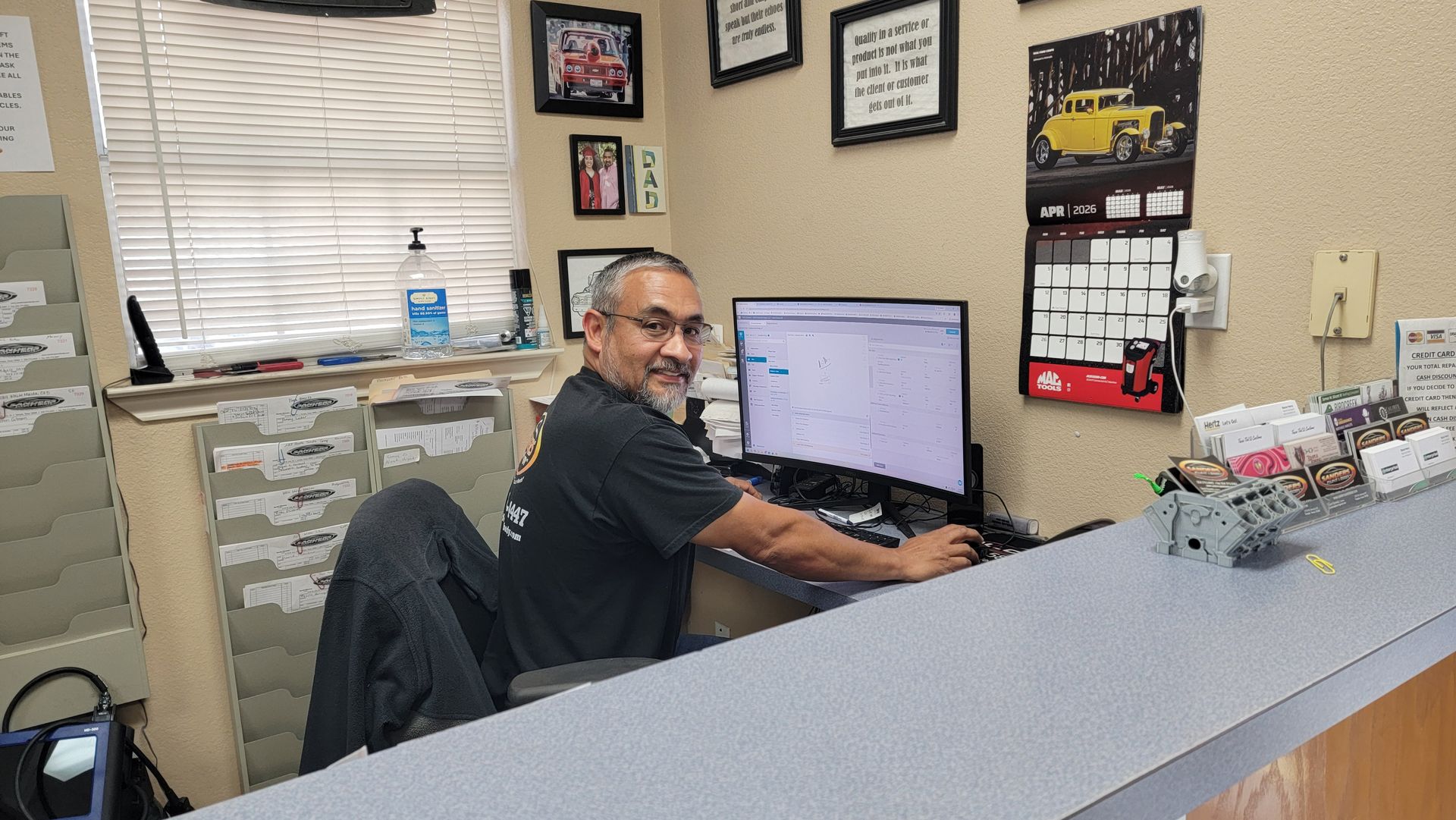 Man sitting at an office desk, smiling at a computer in a small workspace with framed certificates and wall decor.