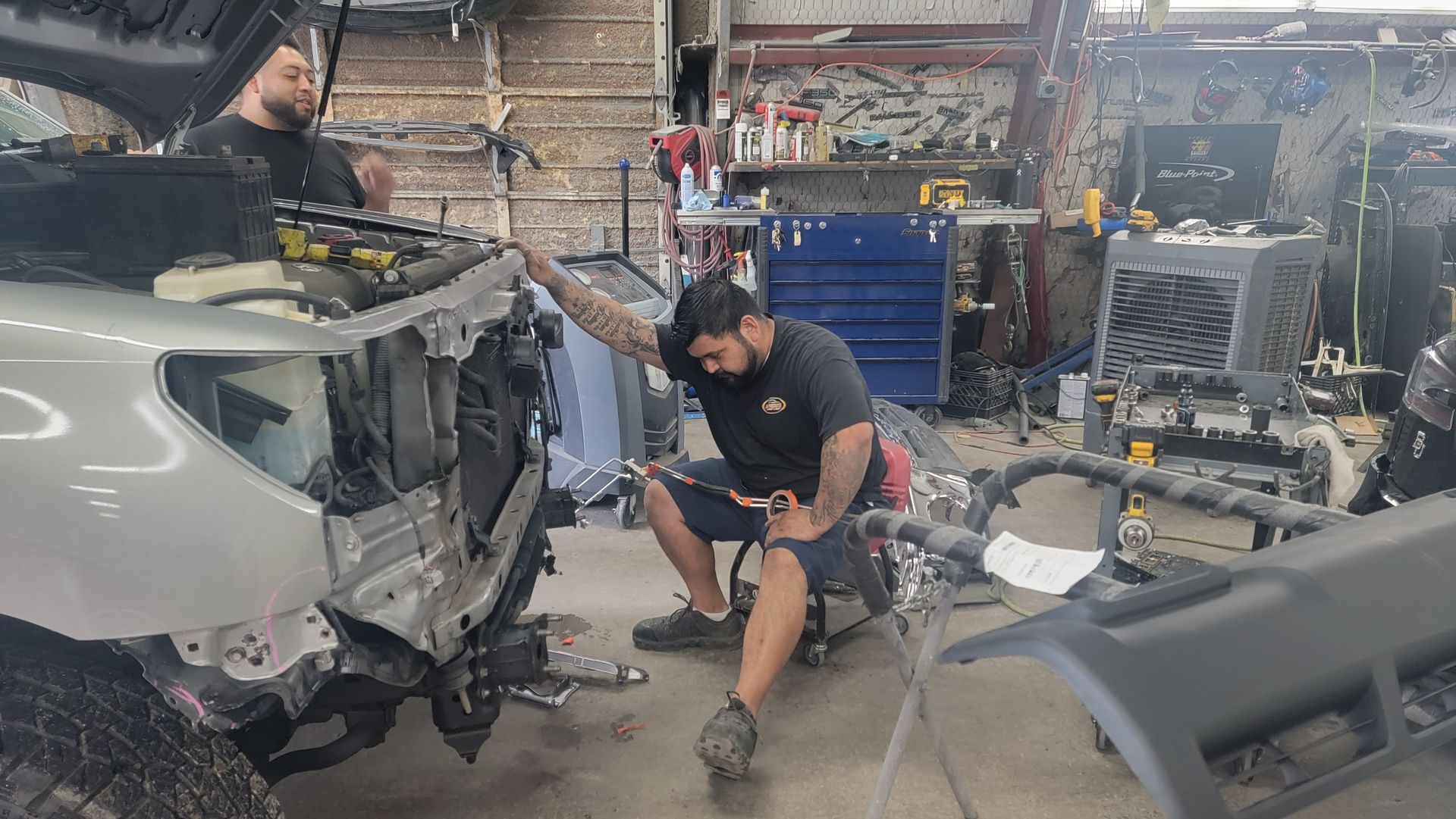 Two mechanics inspect a gray car’s damaged front in a cluttered auto repair garage.
