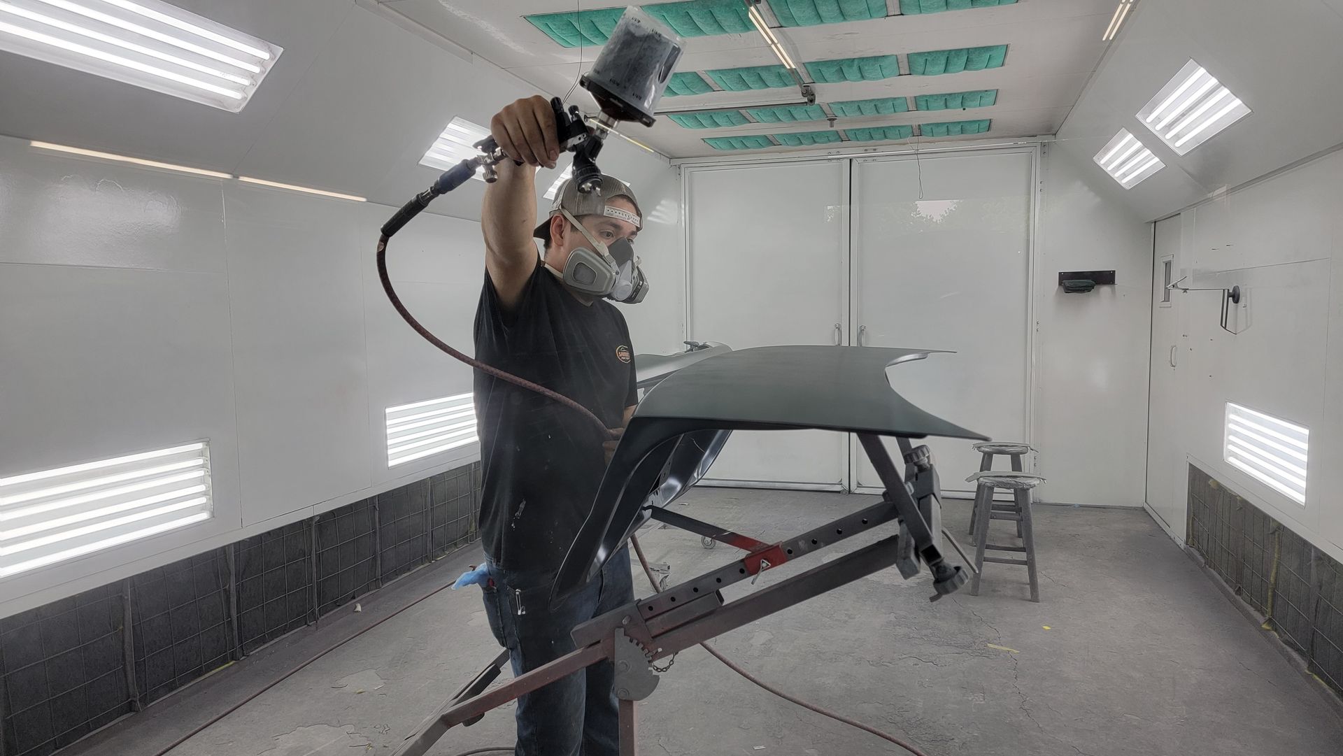 Person sanding a car panel in a spray booth, wearing protective gear and using a power sander.
