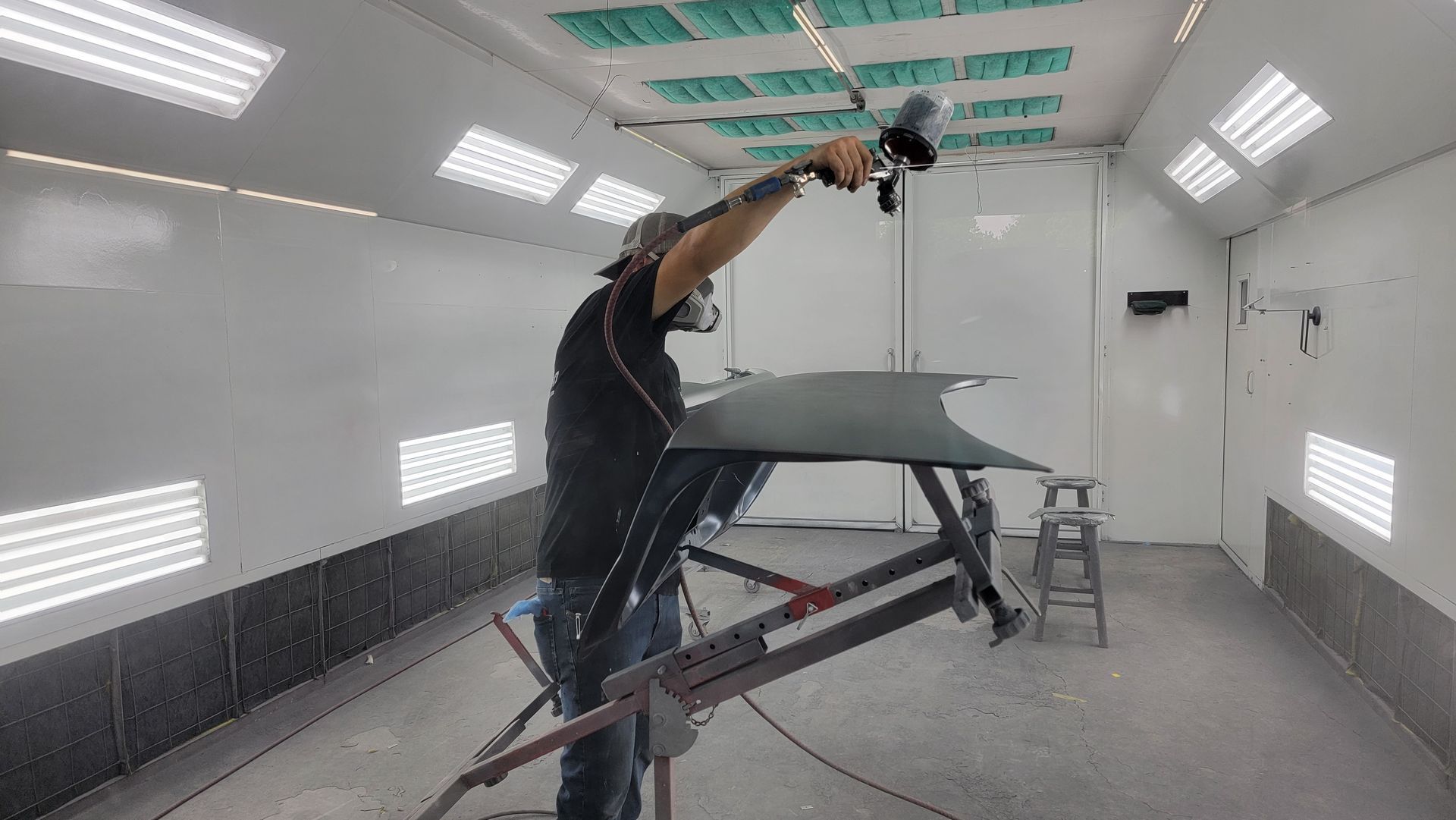 Person sprays a car body in a white paint booth, wearing dark clothes and a face mask.