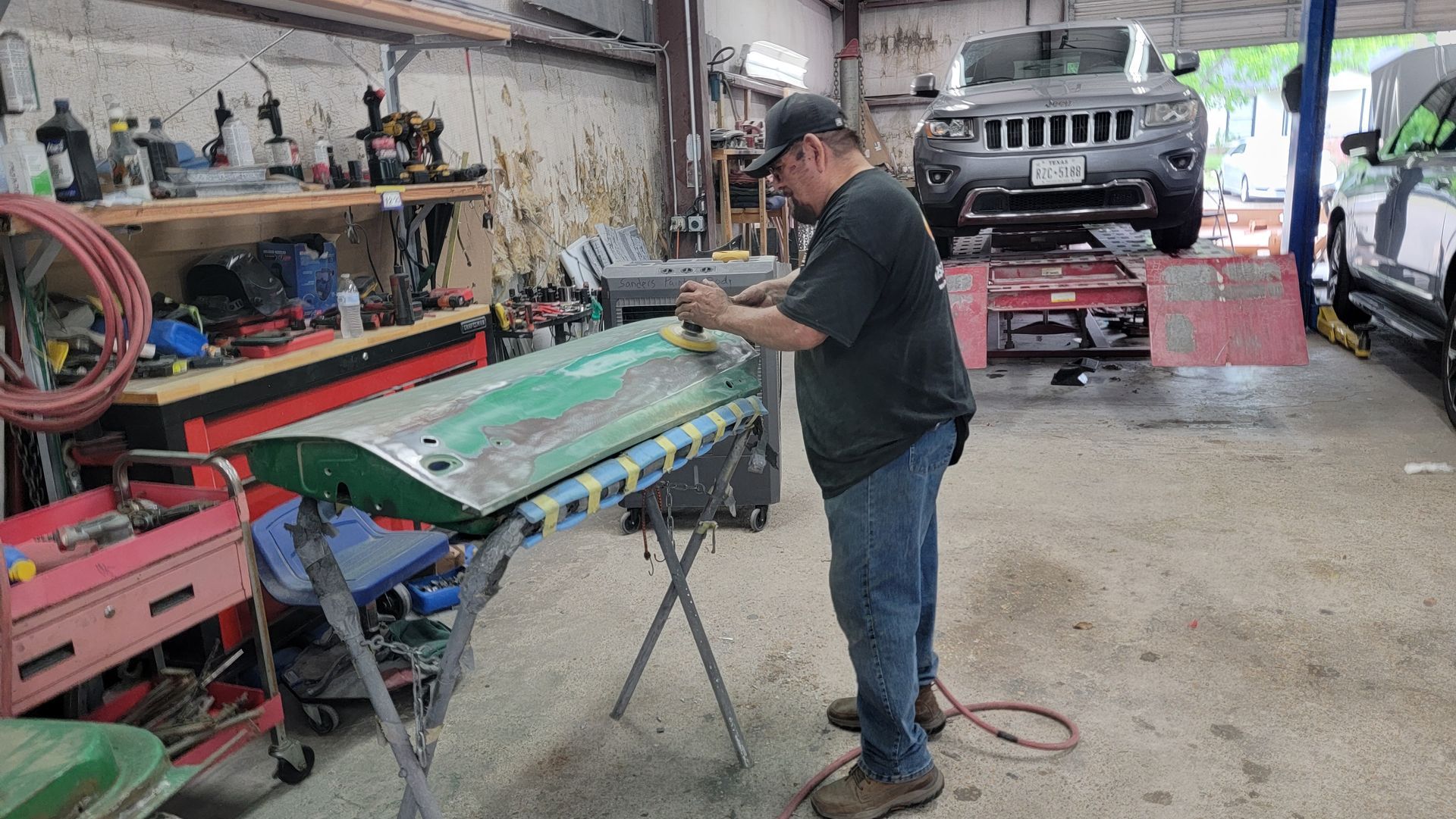 Mechanic working on a green car panel in a cluttered auto repair shop, with a lifted SUV in the background