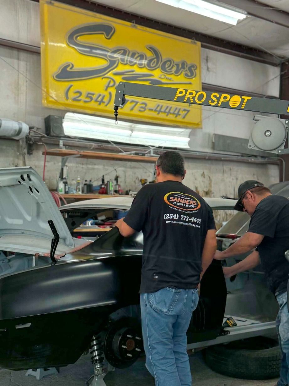 Man working on a car hood in an auto body shop, with tools and a vehicle in the background.