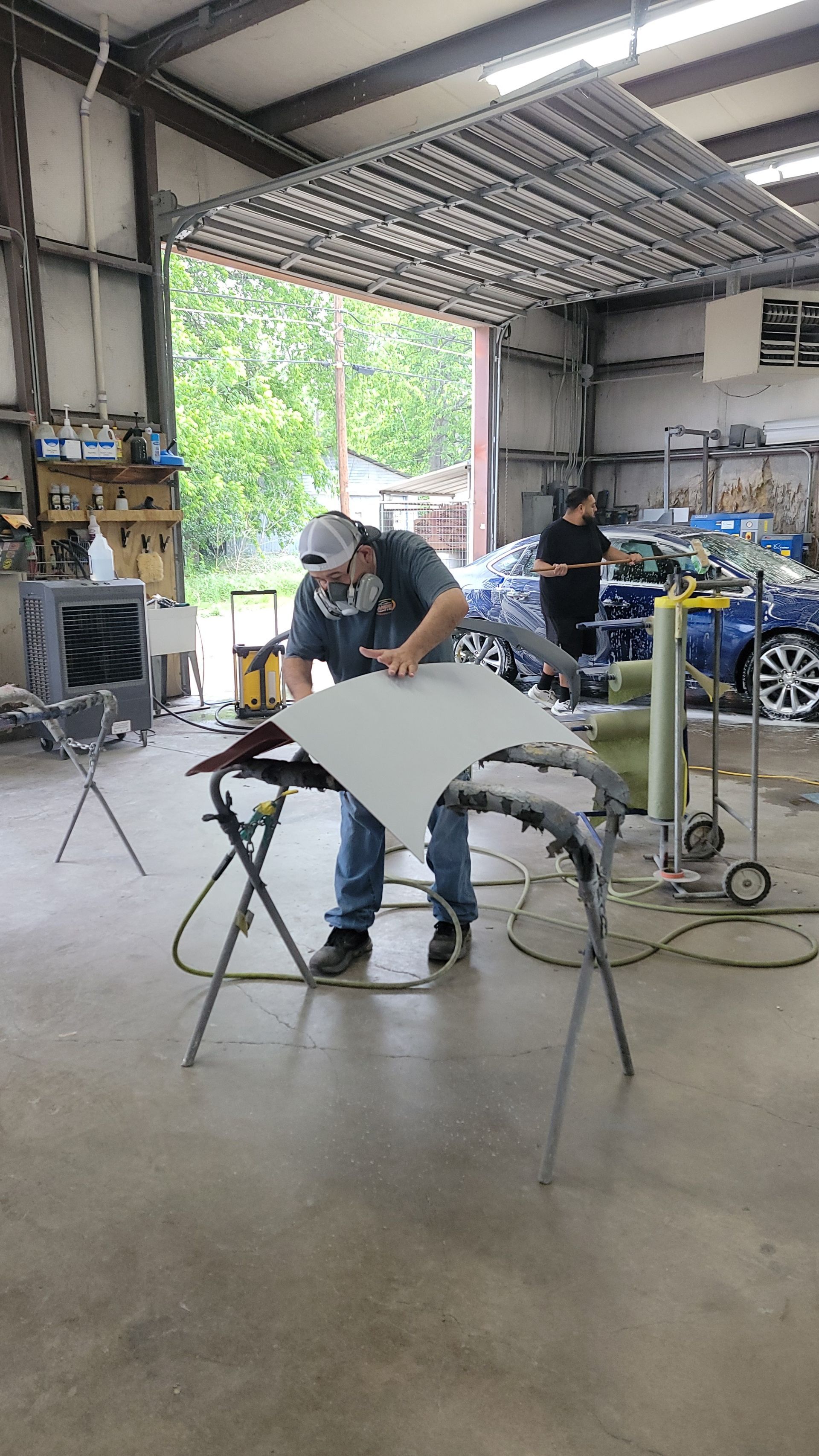 Two people working in a cluttered garage workshop, with a car part on a stand in the foreground.