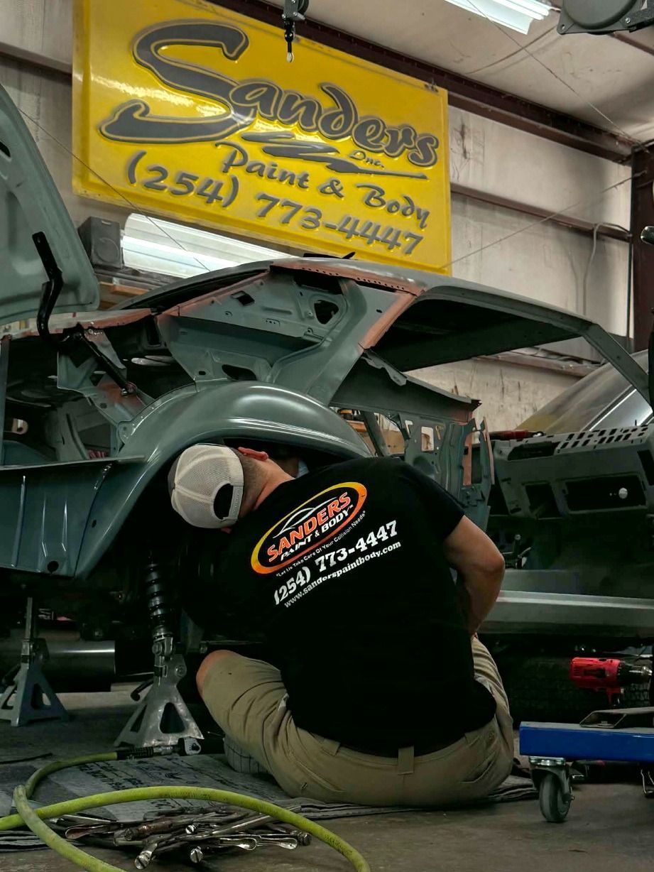 Technician repairing a green vehicle inside a body shop under a yellow sign.