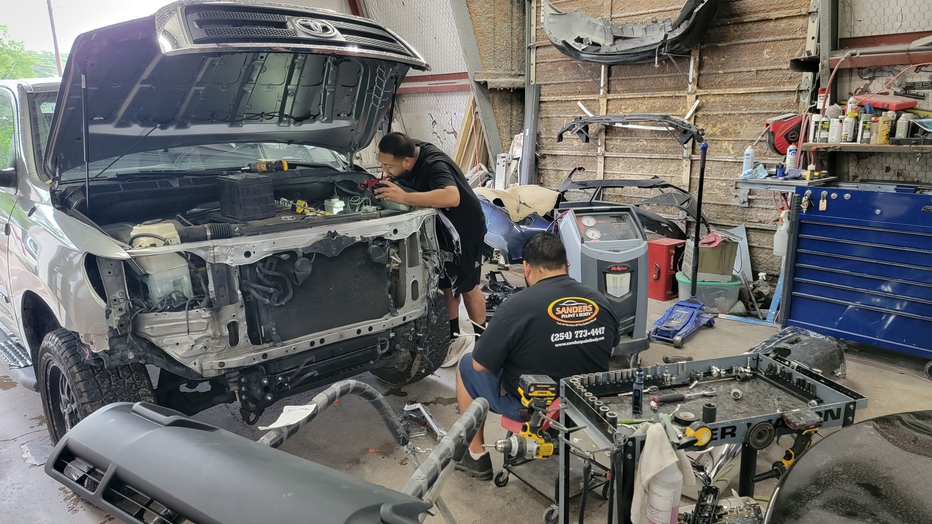 Mechanic repairing a black vehicle in a cluttered auto shop, with tools and parts scattered around