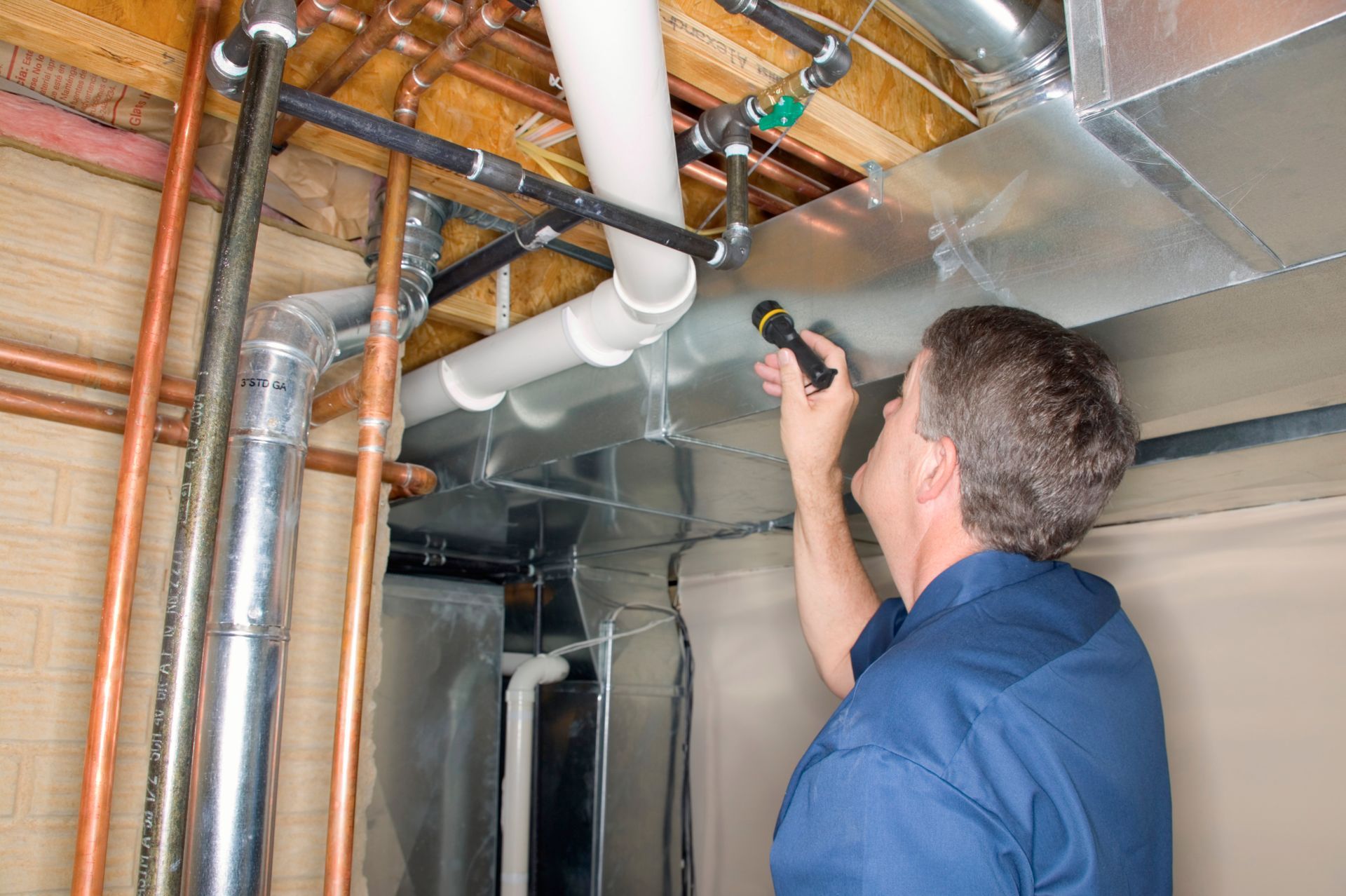 Technician inspecting exposed ceiling pipes with a flashlight in a basement utility area