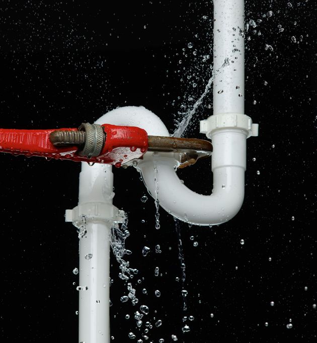 A red pipe wrench grips a white P-trap pipe that is leaking water against a black background.