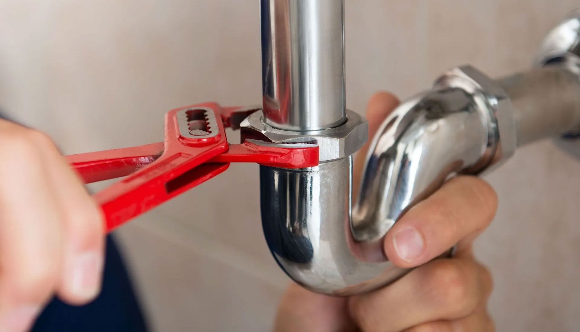 Hands using a red wrench to tighten a chrome plumbing pipe under a sink