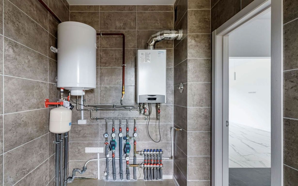 A utility room with a white boiler and water storage tank mounted on tiled walls, featuring exposed plumbing and valves.