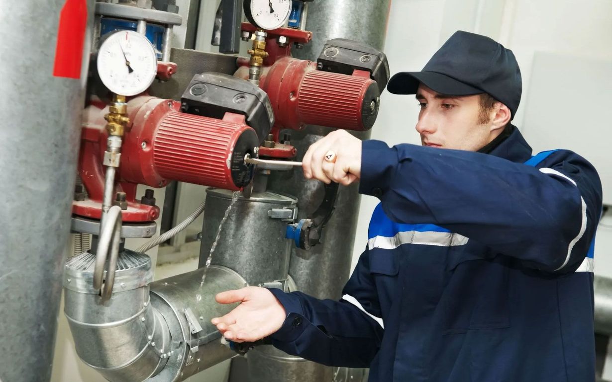 A person in work uniform uses a tool on red industrial pumping equipment in a mechanical room.