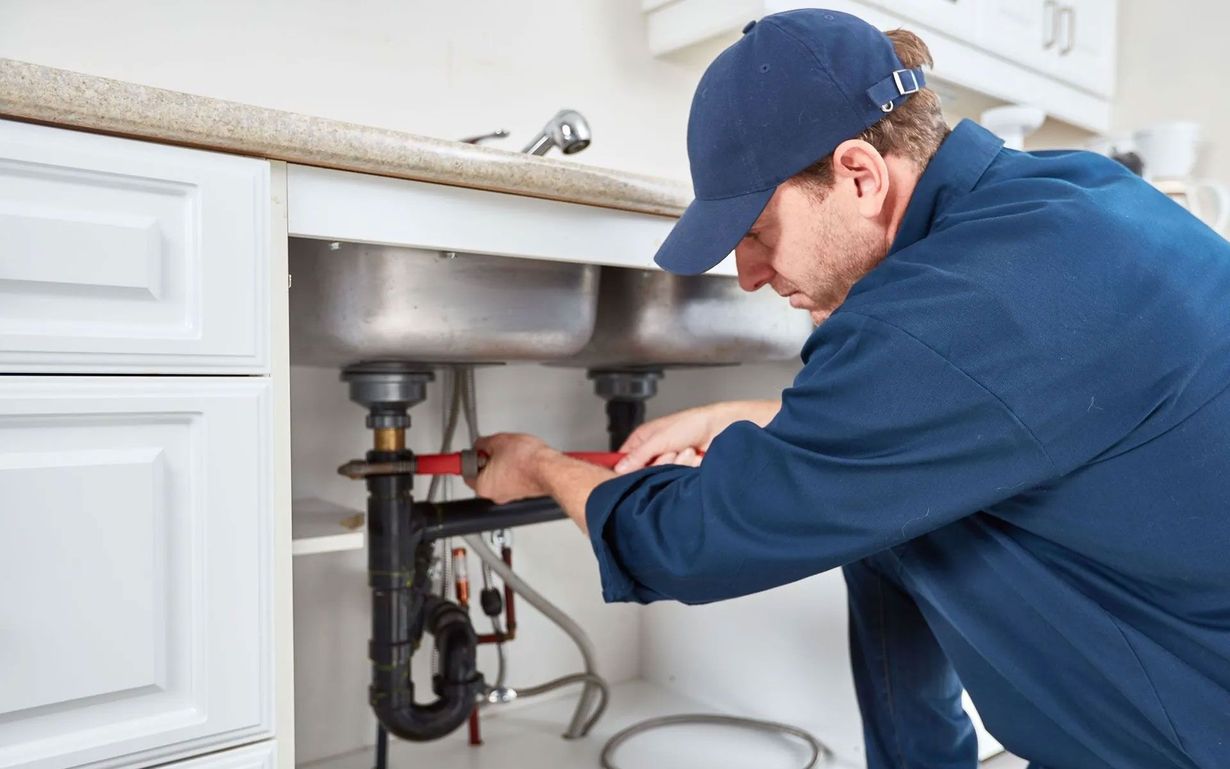 A person wearing a blue cap and work shirt uses a wrench to repair plumbing pipes under a kitchen sink.
