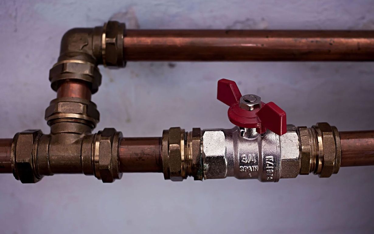 Copper pipes connected by brass fittings with a red-handled shut-off valve, mounted against a plain wall.