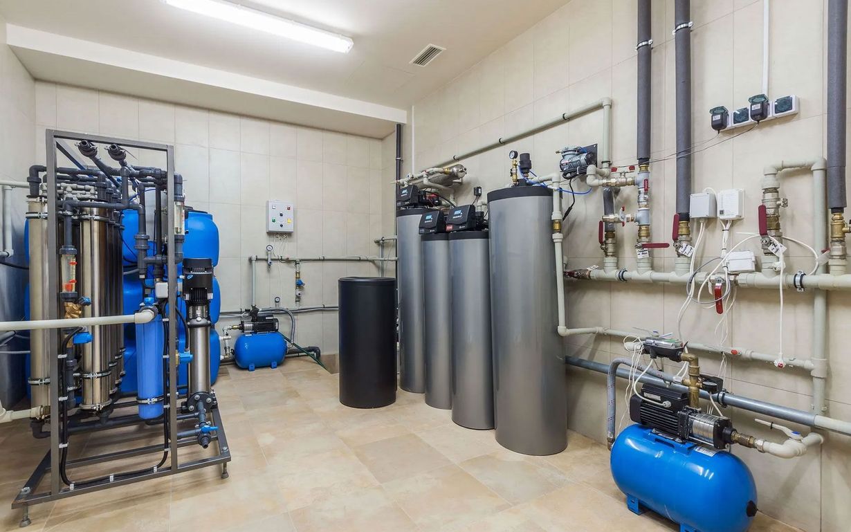 A technical room featuring industrial water filtration equipment, including metal tanks, blue pumps, and piping.