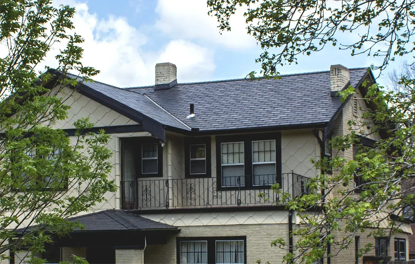 A two-story stucco house with a dark roof and black window trim, partially framed by green tree branches.