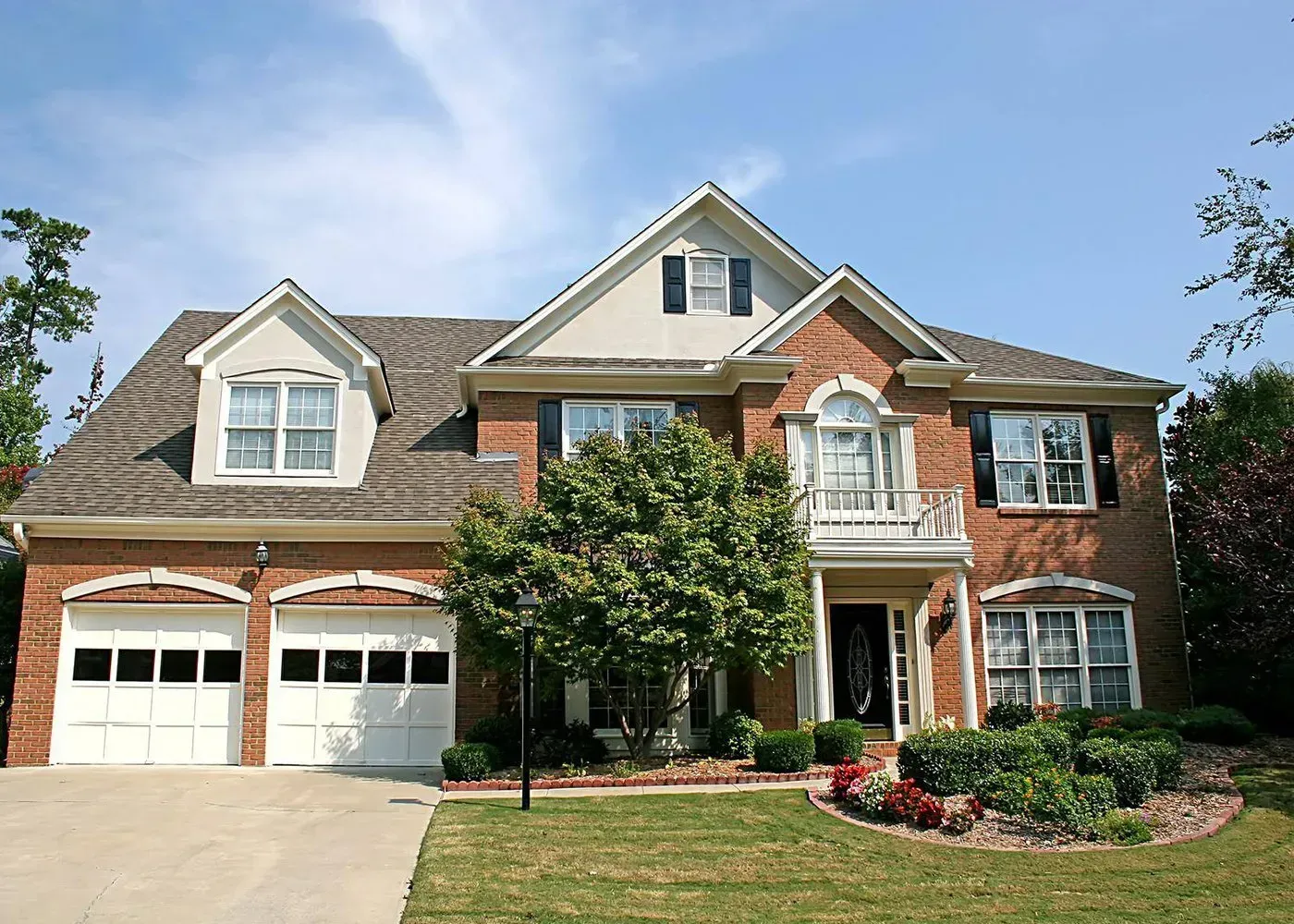 A two-story, red brick house with a front-facing garage, a small upper balcony, and a green tree in the front yard.