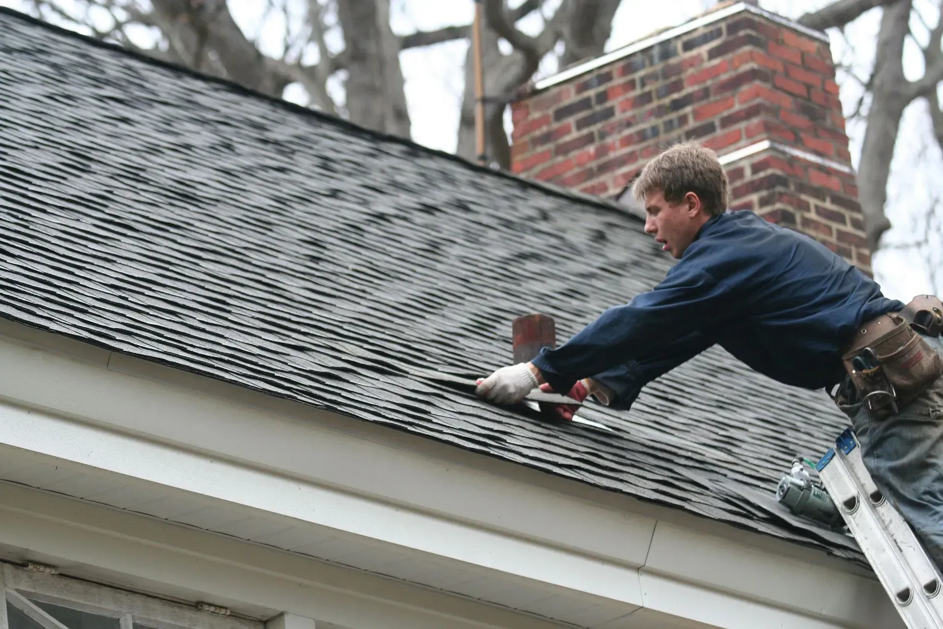 A person on a ladder repairs shingles on a residential roof near a brick chimney.