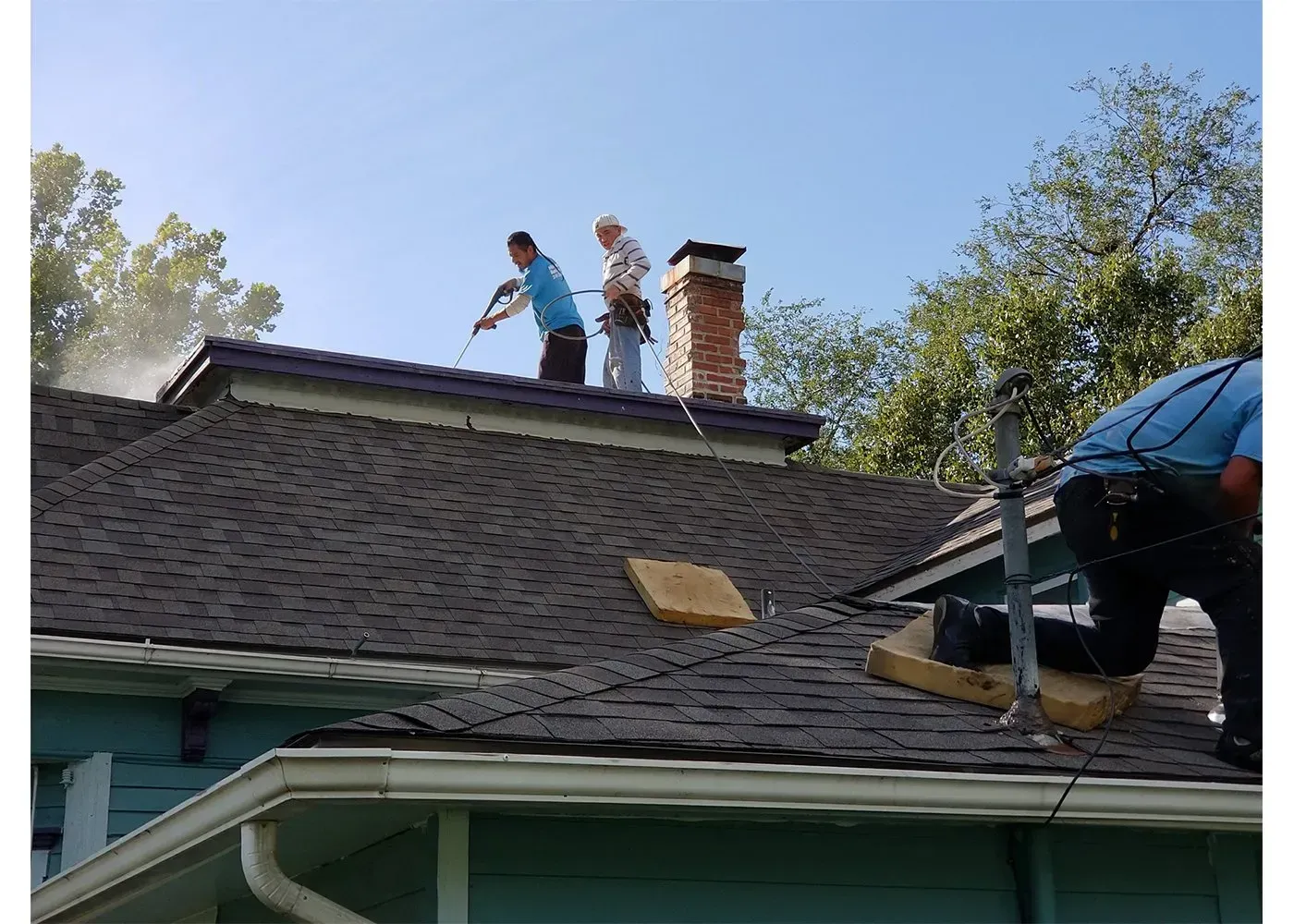 Two workers pressure-wash a dark shingled roof near a brick chimney on a sunny day.