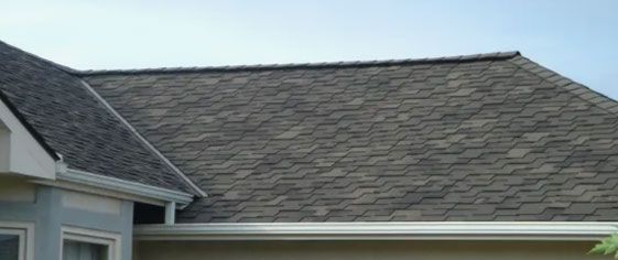 The brown asphalt shingle roof of a house with a white gutter system against a pale blue sky.