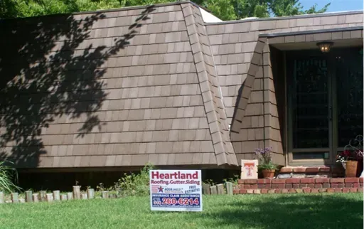 A brown shingled home with a slanted roof and a yard sign for Heartland Roofing, Gutter, and Siding in the grass.
