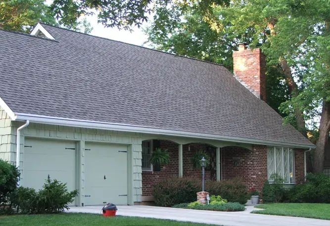 A green house with a brick facade, a two-car garage, and a steeply pitched shingled roof under mature trees.
